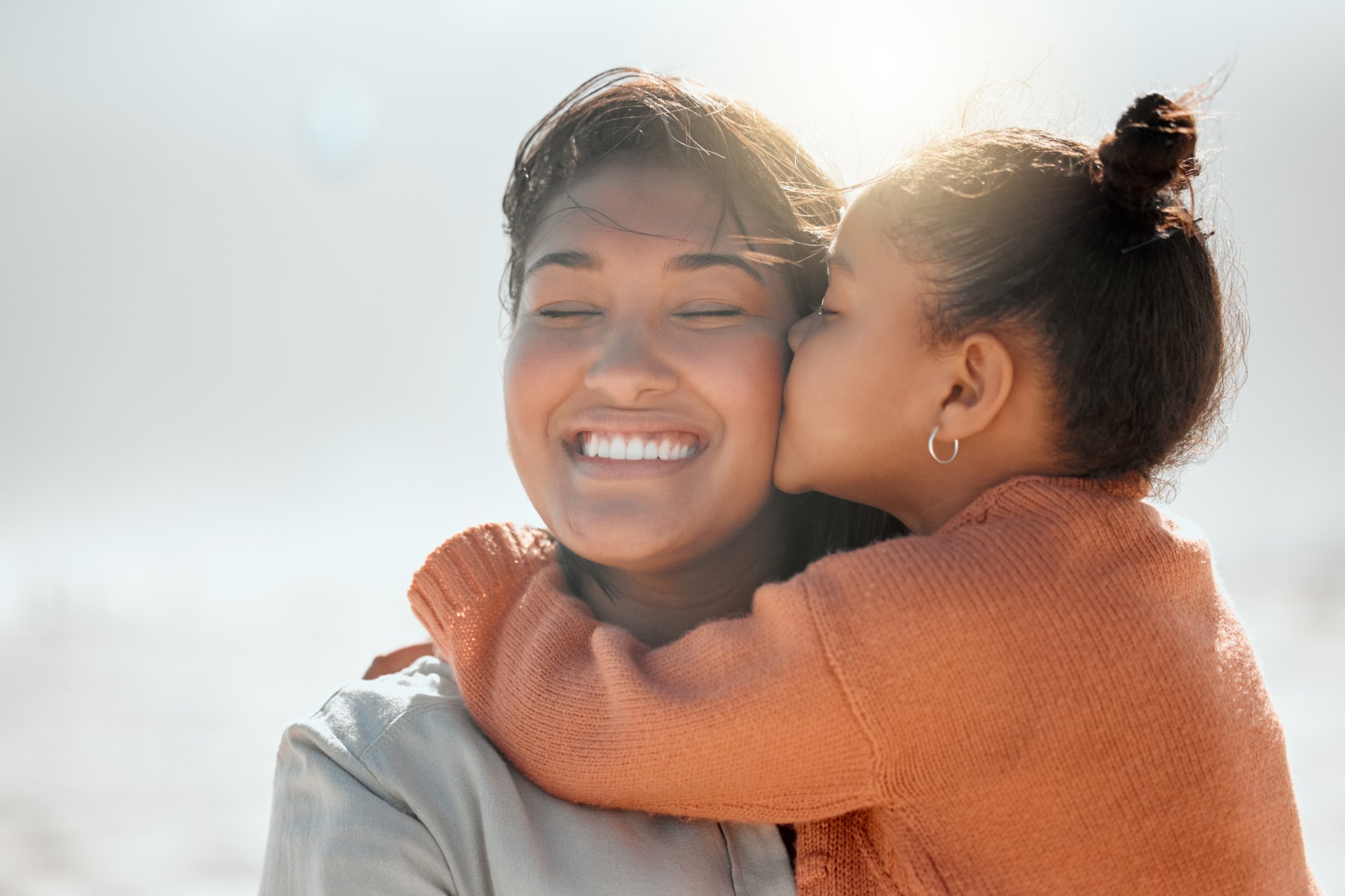 Adorable mixed race girl kisses her mother on the cheek and outs her arms around her while at the b Adorable mixed race girl kisses her mother on the cheek and outs her arms around her while at the b.